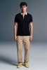 Full-length front view of a man standing and facing forward, wearing a open-collar black polo shirt, khaki trousers, and yellow shoes.