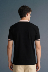 Full-length rear view of a man standing straight, wearing a black short-sleeved top with a light-colored collar and patterned sleeve trims, paired with beige trousers.