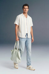 Man wearing a white striped shirt and light blue jeans, holding a light green bag against a gray background