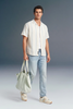 Man wearing a white striped shirt and light blue jeans, holding a light green bag against a gray background