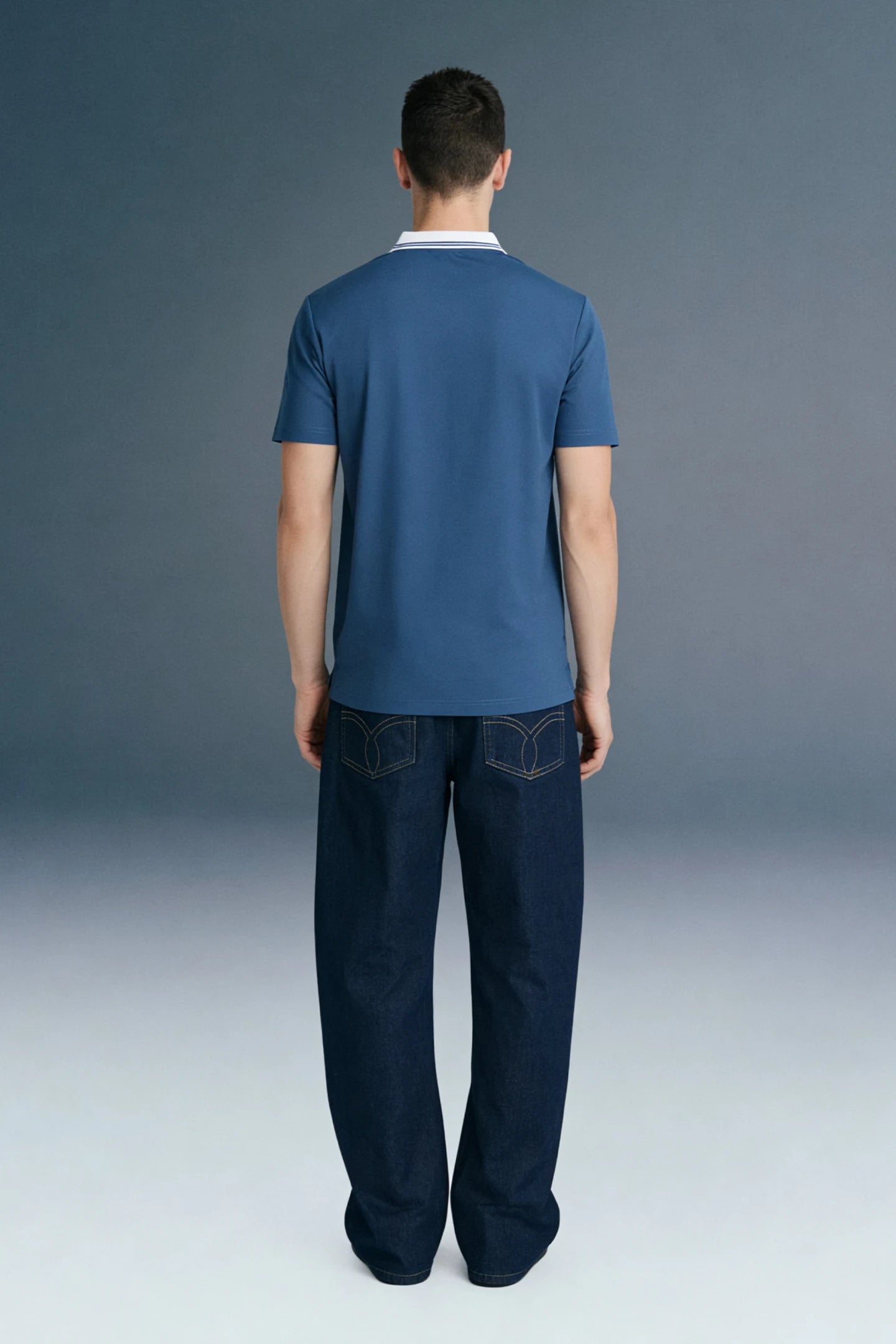 Full-length rear view of a man standing straight, wearing a slate blue polo shirt with a white collar and dark navy blue trousers.