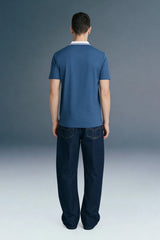 Full-length rear view of a man standing straight, wearing a slate blue polo shirt with a white collar and dark navy blue trousers.