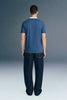 Full-length rear view of a man standing straight, wearing a slate blue polo shirt with a white collar and dark navy blue trousers.
