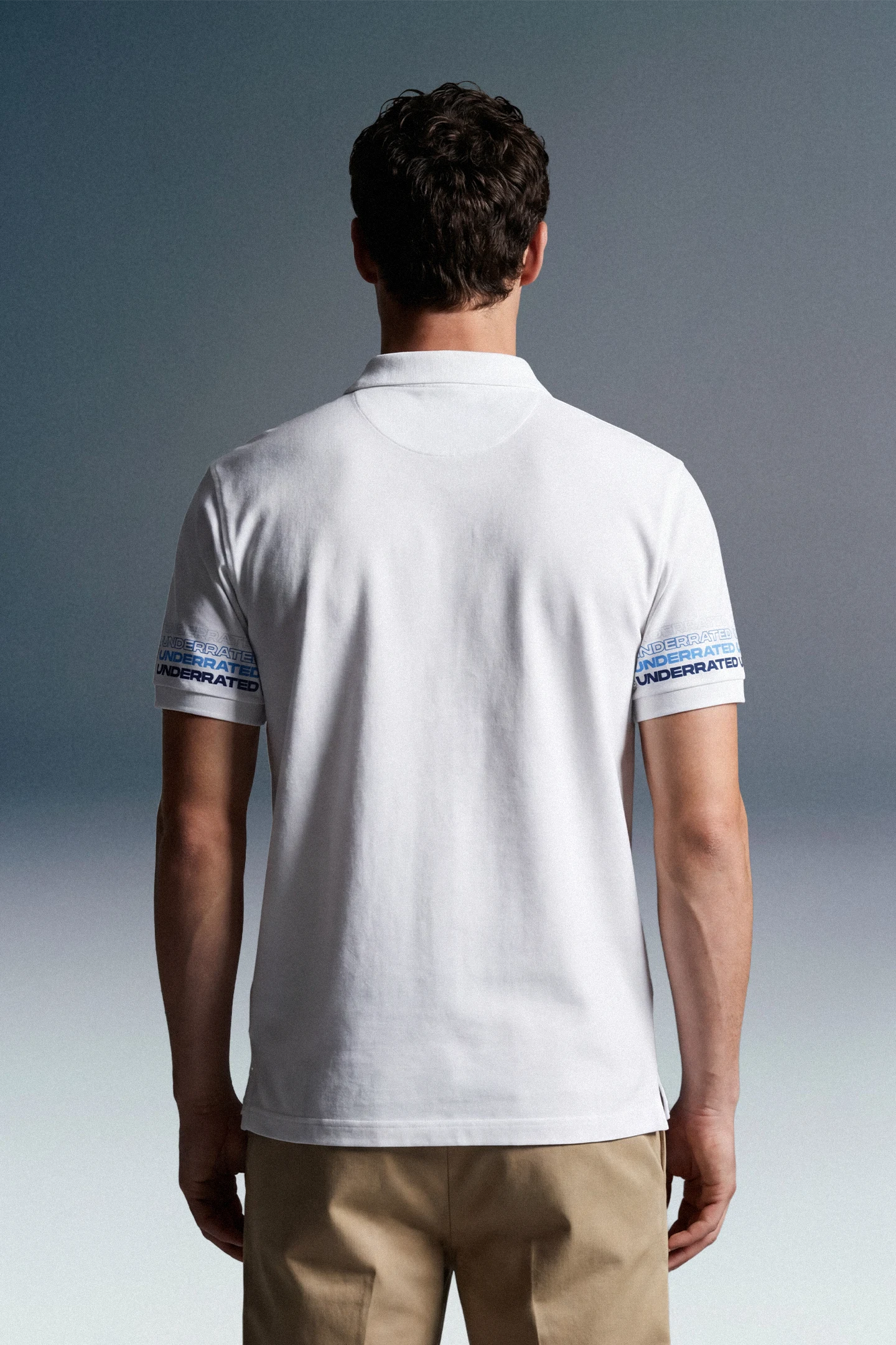 Rear view of a man with dark, wavy hair wearing a white polo shirt and khaki trousers. The short sleeves of the polo feature a repeating blue graphic pattern or text running horizontally around the cuffs.
