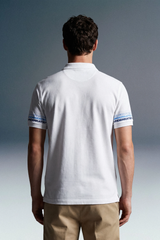 Rear view of a man with dark, wavy hair wearing a white polo shirt and khaki trousers. The short sleeves of the polo feature a repeating blue graphic pattern or text running horizontally around the cuffs.