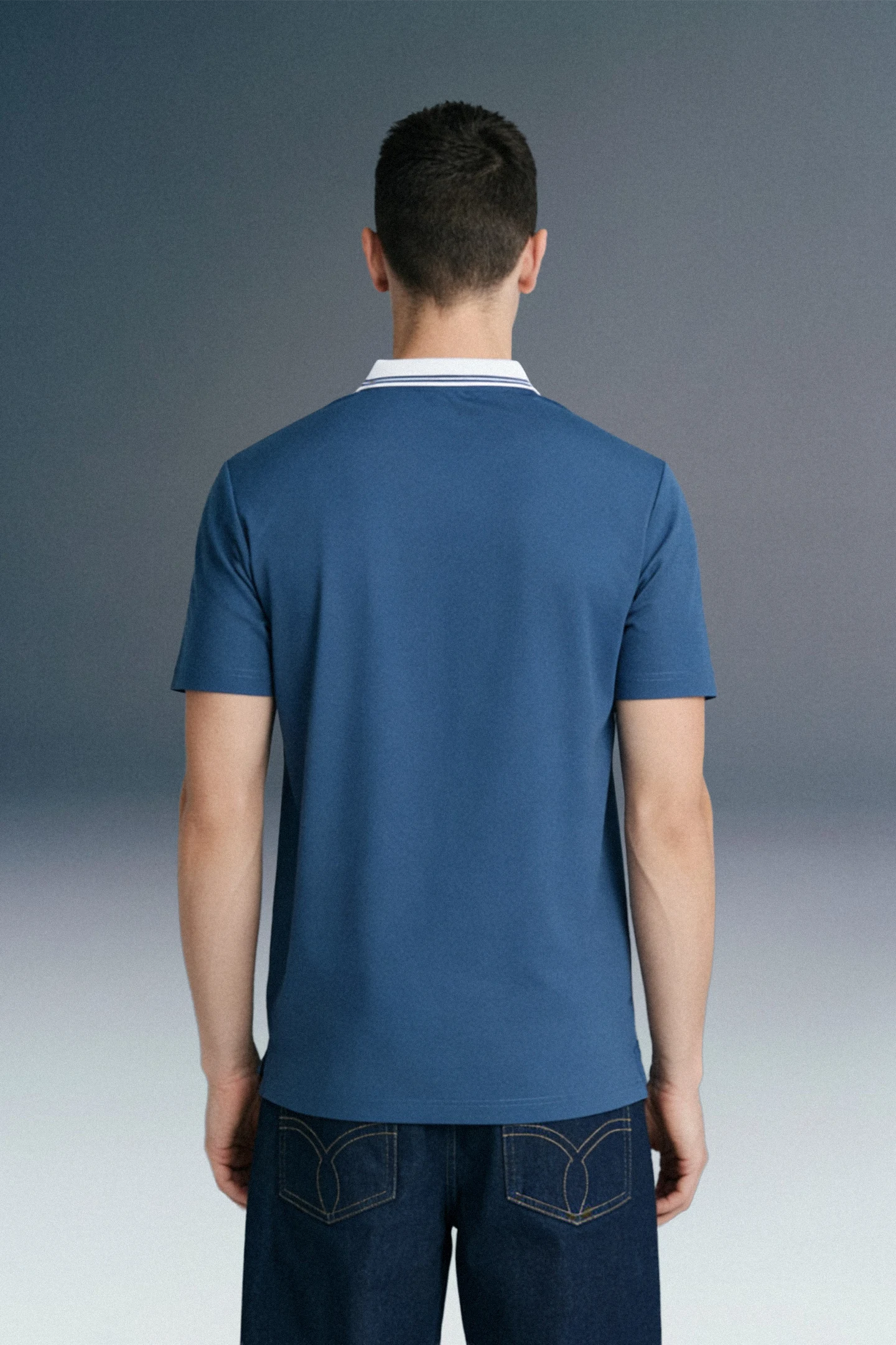 Rear view from the waist up of a man wearing a slate blue polo shirt with a contrasting white collar.